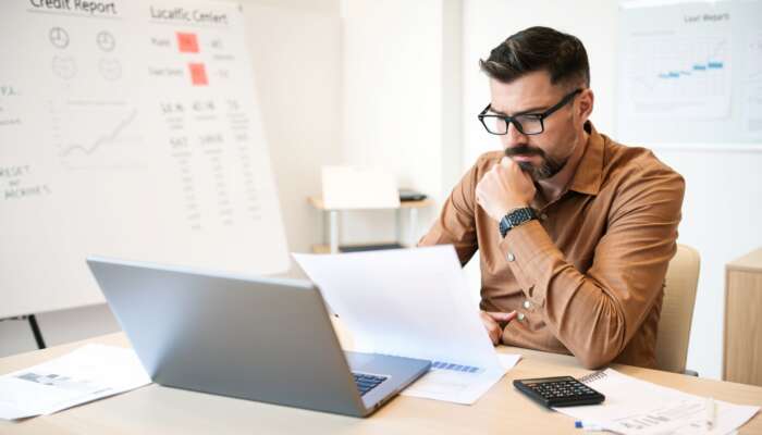 Business owner reviewing a digital credit report on a laptop in an office, surrounded by financial documents, a calculator, and a notepad, showing determination in monitoring credit health.